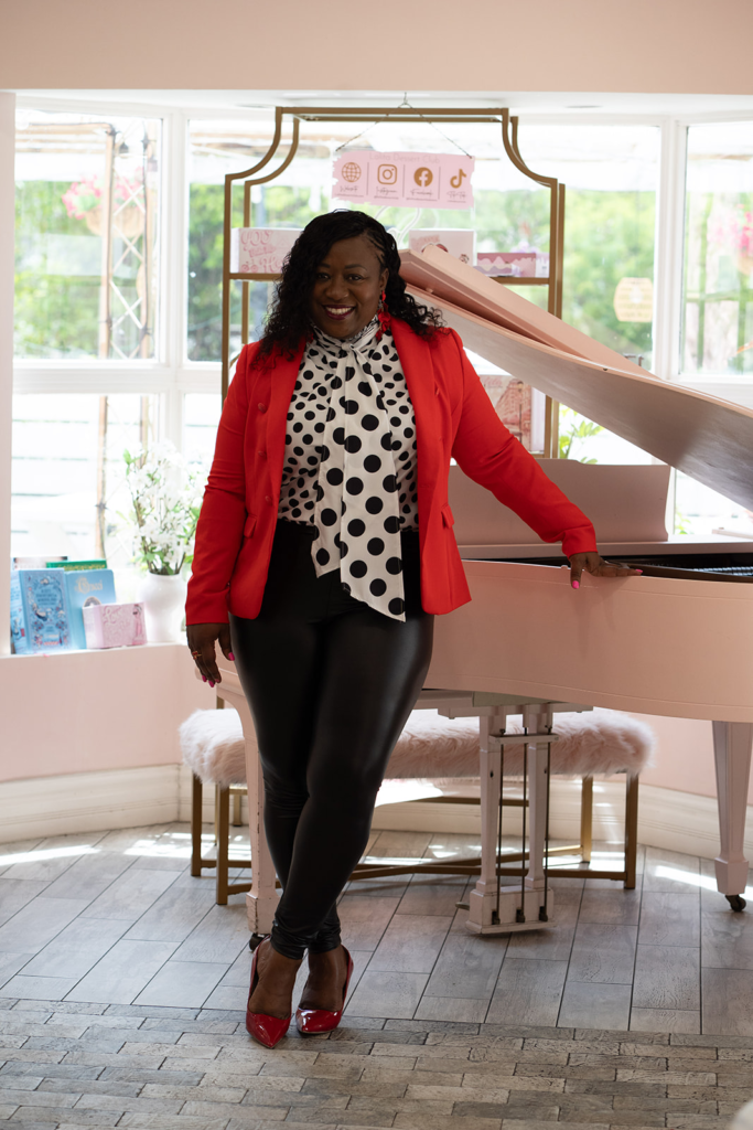 Audrey Morison standing confidently in a bright space beside a white piano, wearing a red blazer and polka-dot blouse